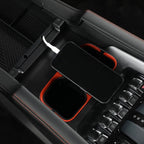 Black center console organizer with orange accents and a smartphone charging via white cable.