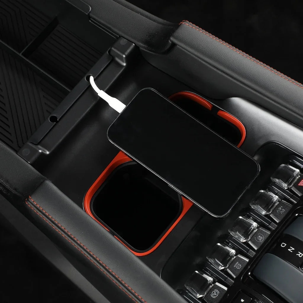 Black center console organizer with orange accents and a smartphone charging via white cable.