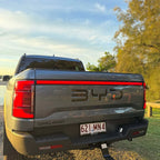 Rear view of a grey BYD Shark pickup truck parked outdoors near water under a clear sky.