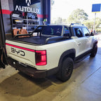 White BYD Shark 6 pickup truck with black roll bar and matte black tonneau cover in a garage.