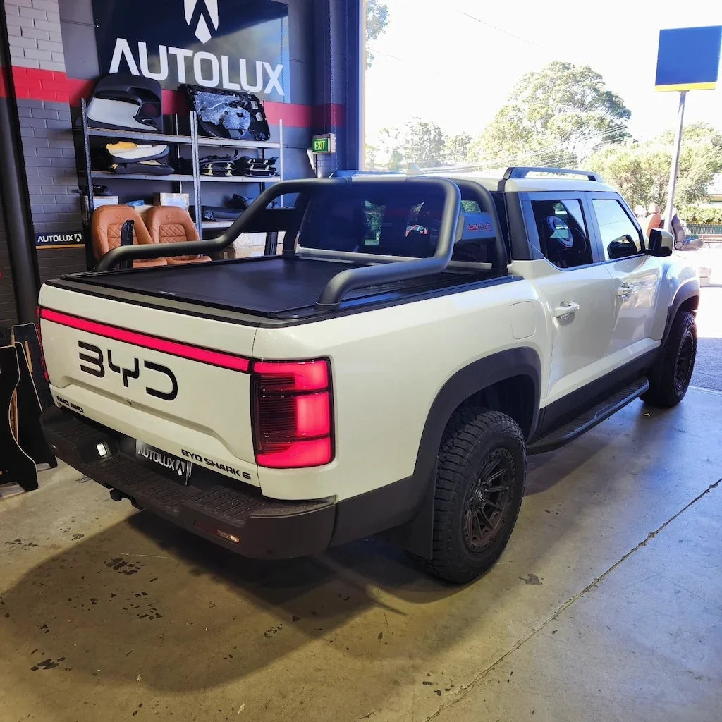 White BYD Shark 6 pickup truck with black roll bar and matte black tonneau cover in a garage.