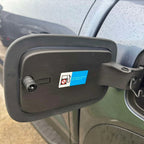 Black matte plastic side mirror housing with a white fuel type warning sticker on a grey car.