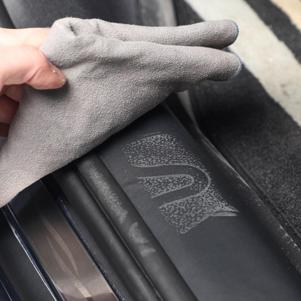 A person uses a grey microfiber cloth to clean a black protective car sill plate.