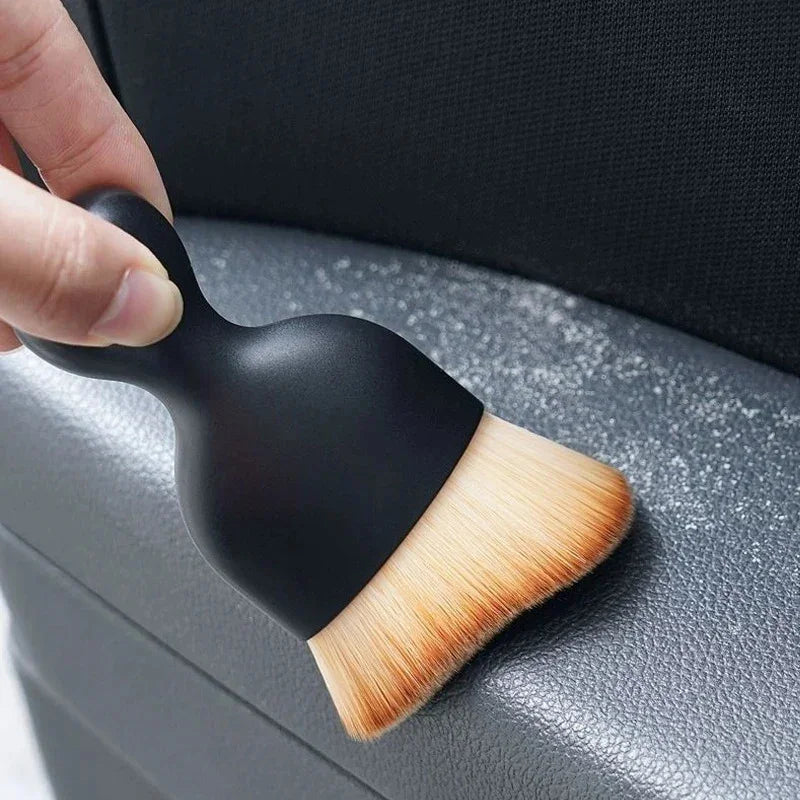 Black detailing brush with soft tan bristles cleaning dust from a grey car interior surface.