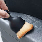Black detailing brush with soft tan bristles cleaning dust from a grey car interior surface.