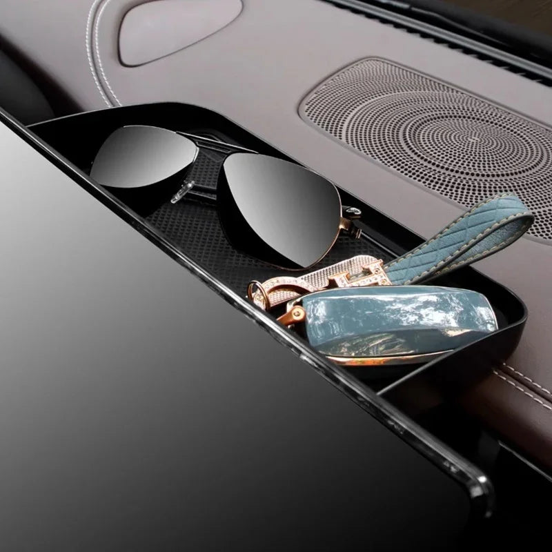 Black leather car center console organizer tray holding dark sunglasses and a blue quilted accessory.