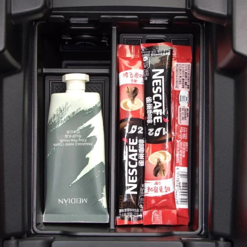 Black car interior storage compartment containing a white hand cream tube and coffee sachets.