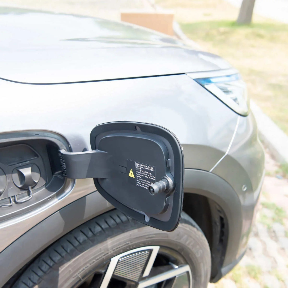 Close-up of an open grey charging port cover on a modern electric vehicle parked outdoors.