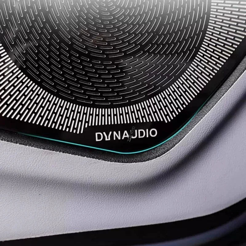Close-up of a black Dynaudio speaker grille with a sleek metallic texture on grey upholstery.