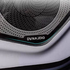 Close-up of a black Dynaudio speaker grille with a sleek metallic texture on grey upholstery.