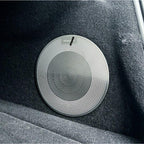 Silver metallic circular speaker grille with a textured concentric pattern on dark grey car interior fabric.