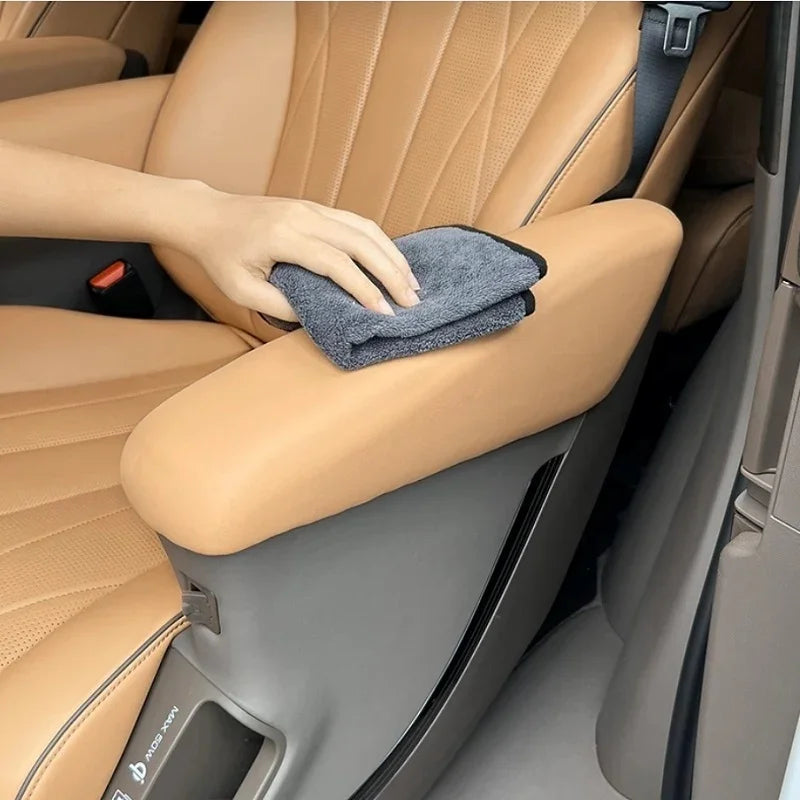 A hand using a grey microfiber cloth to clean a tan leather car armrest and seat interior.