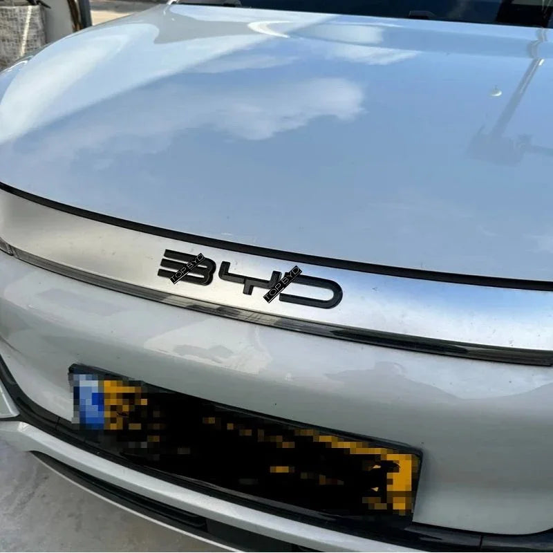 Close-up of a silver BYD car hood featuring a bold, black 3D logo emblem.
