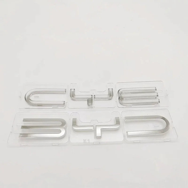 Silver metallic BYD car emblems in transparent plastic protective packaging.