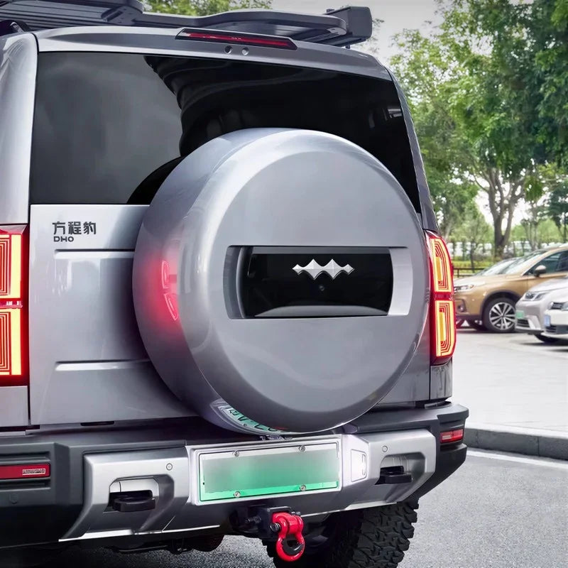 Silver metallic hard-shell spare tire cover with a sleek black decorative emblem on a grey SUV.