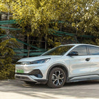 Sleek white BYD electric SUV parked outdoors near lush green trees and a brick wall.