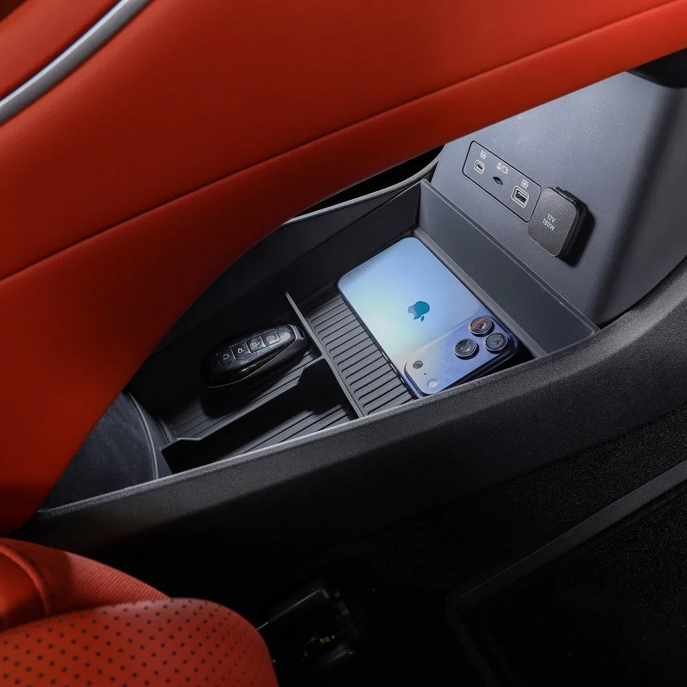 Close-up of a black plastic car storage console with a smartphone and key fob near red leather seats.