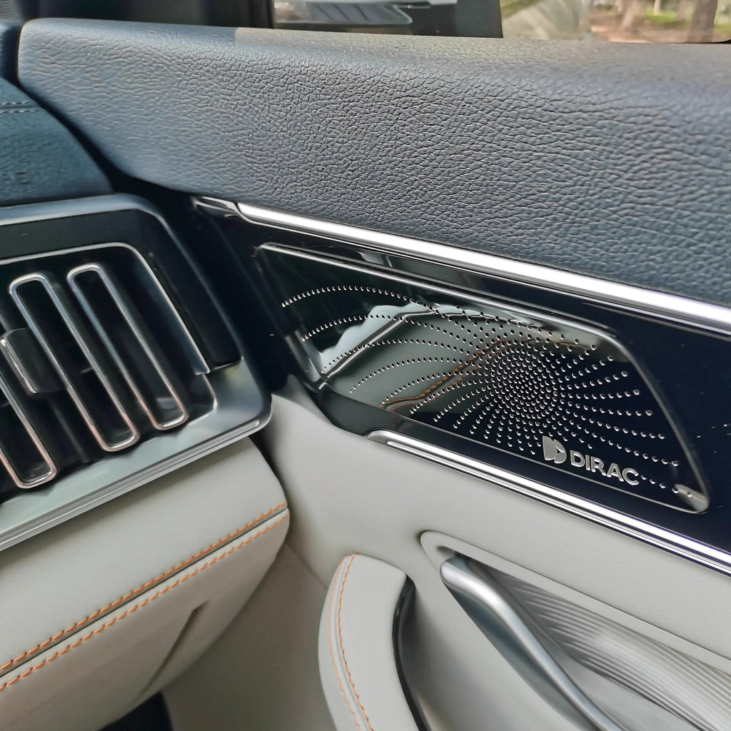 Black Dirac premium speaker grille with a polished metallic finish on a grey leather car interior.