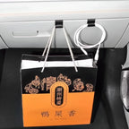 A black and orange paper gift bag with white rope handles hanging from a car seat hook next to a white charging cable.