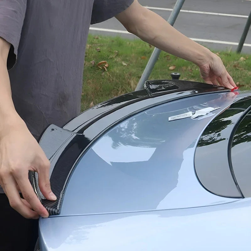 A person installing a sleek black carbon fiber rear spoiler on a light blue BYD car.