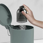 Hand pouring white granules from a transparent black plastic dispenser into a matte grey bin.