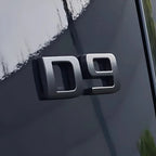 Silver metallic D9 emblem mounted on a sleek dark grey car body surface.