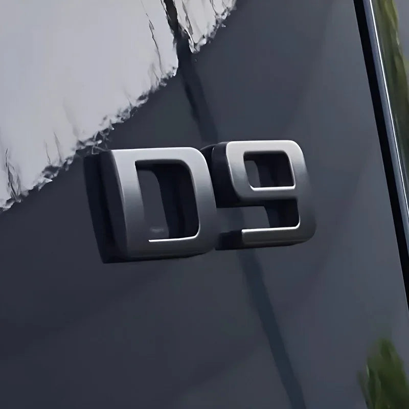 Silver metallic D9 emblem mounted on a sleek dark grey car body surface.