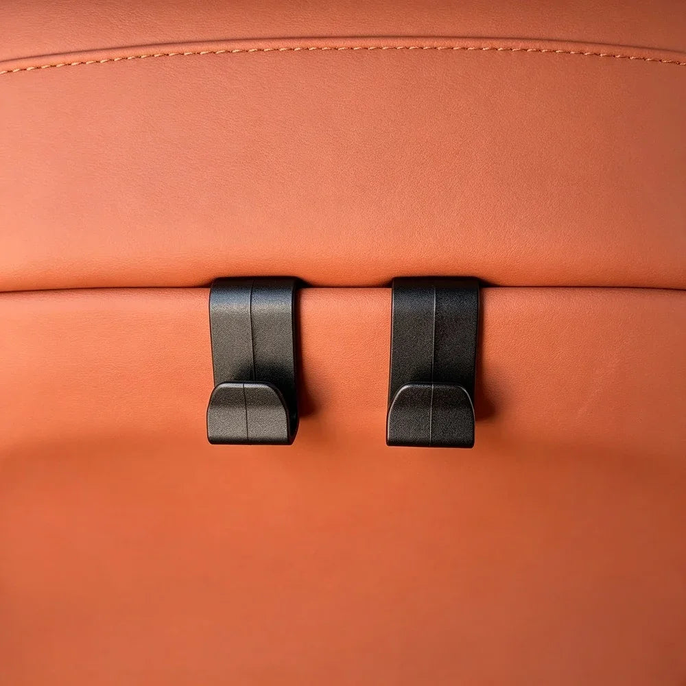 Two black matte plastic seat hooks mounted on premium tan leather car upholstery.