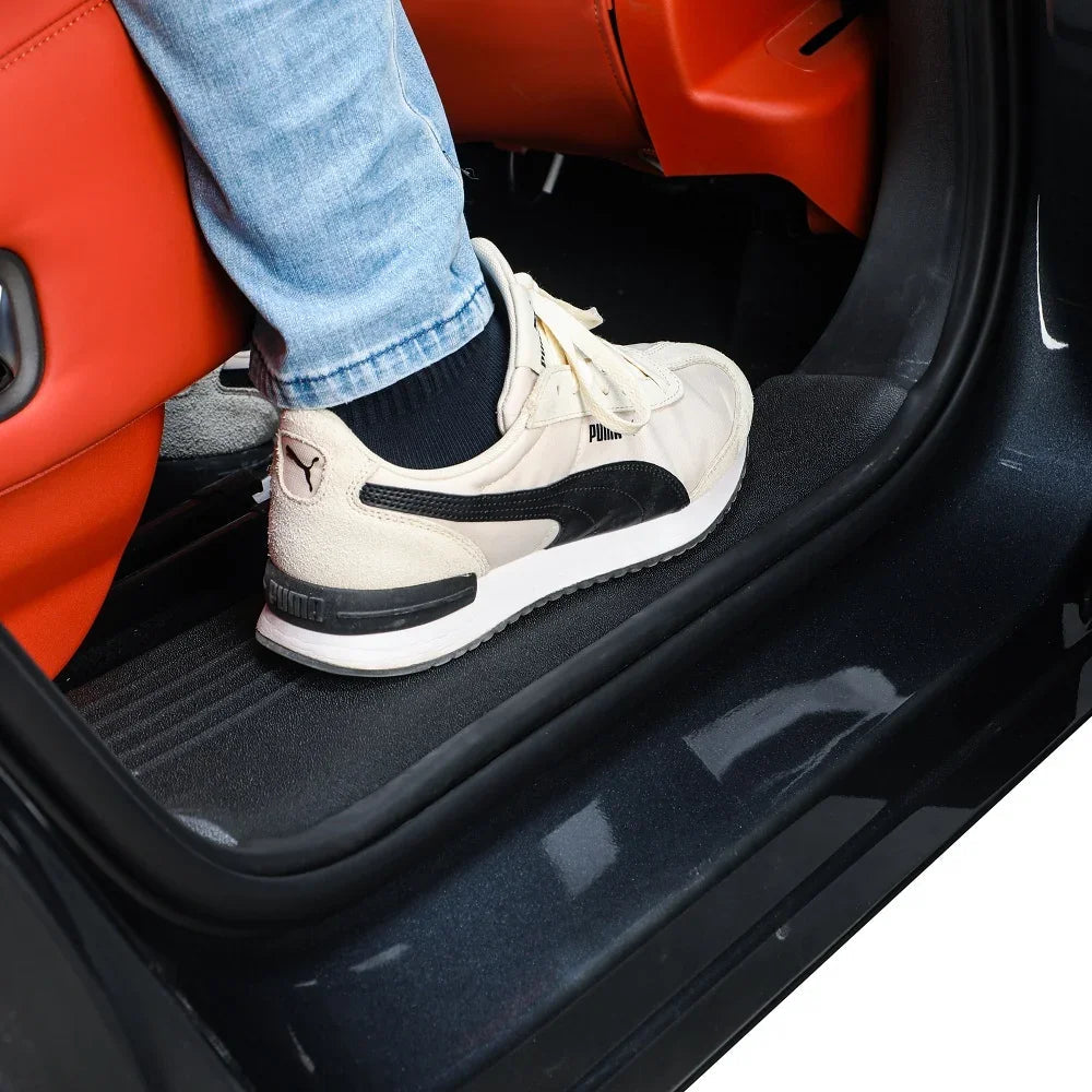 A person wearing cream Puma sneakers and blue jeans stepping onto a black rubber car floor mat.