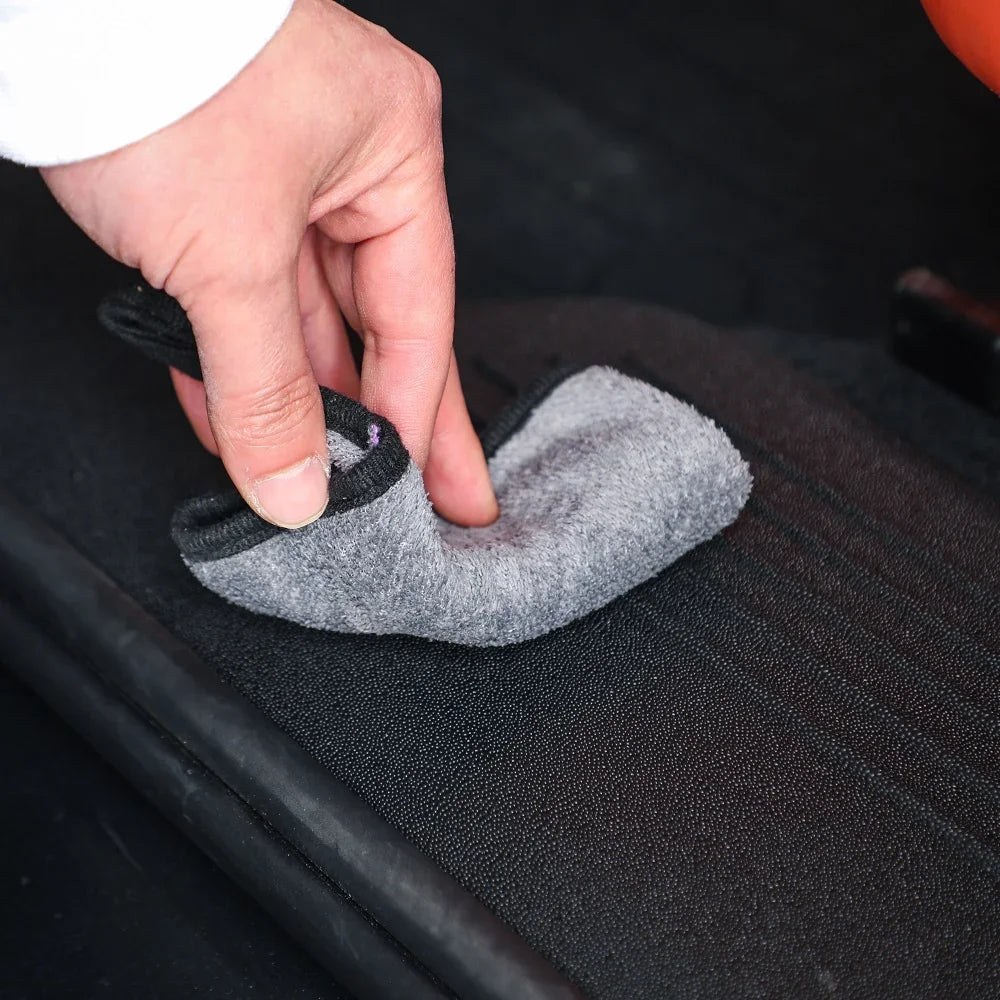 A hand using a grey microfiber cloth to clean a black textured car interior surface.