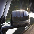 Close-up of a car side mirror cover featuring a grey carbon fiber texture and integrated LED light strip.