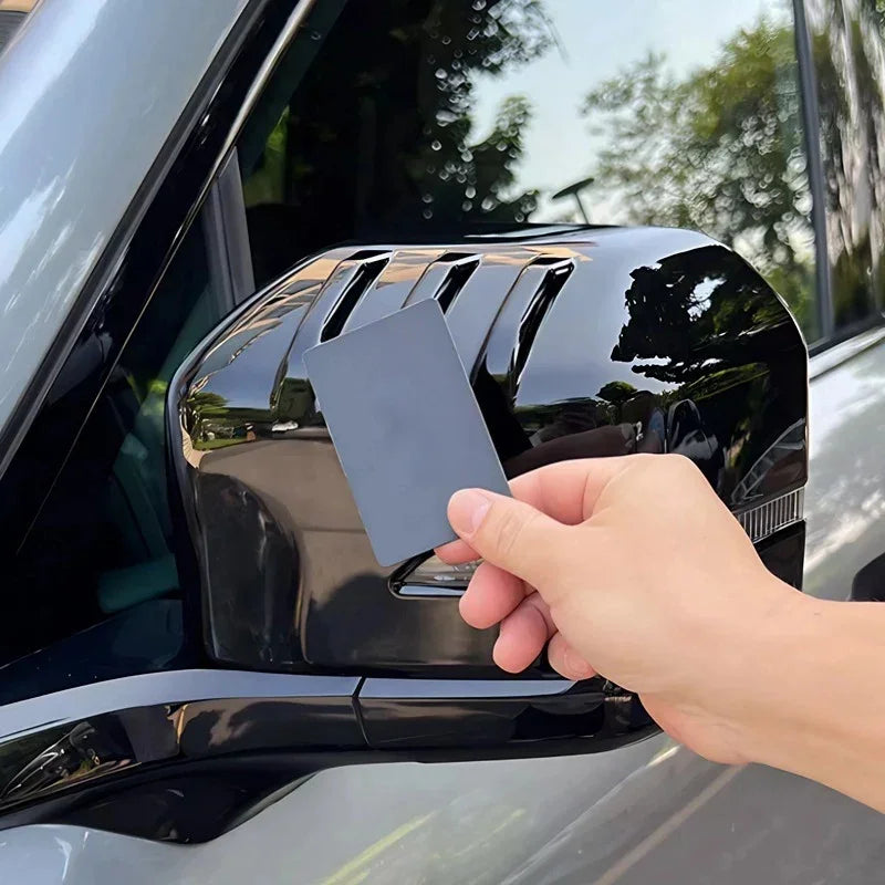 Glossy black side mirror cover being applied with a protective film on a grey car.