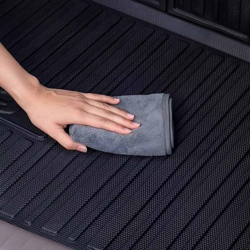 Hand wiping a black textured TPE car floor mat with a grey microfiber cloth.