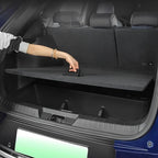 A person's hand lifting a black fabric cargo privacy cover in the trunk of a blue car.