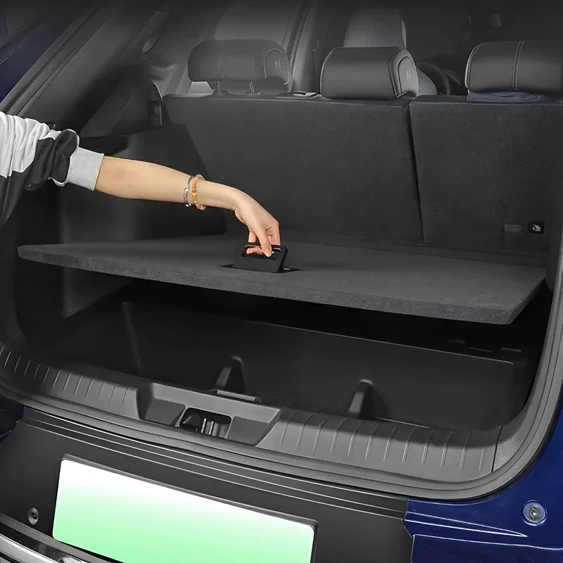 A person's hand lifting a black fabric cargo privacy cover in the trunk of a blue car.
