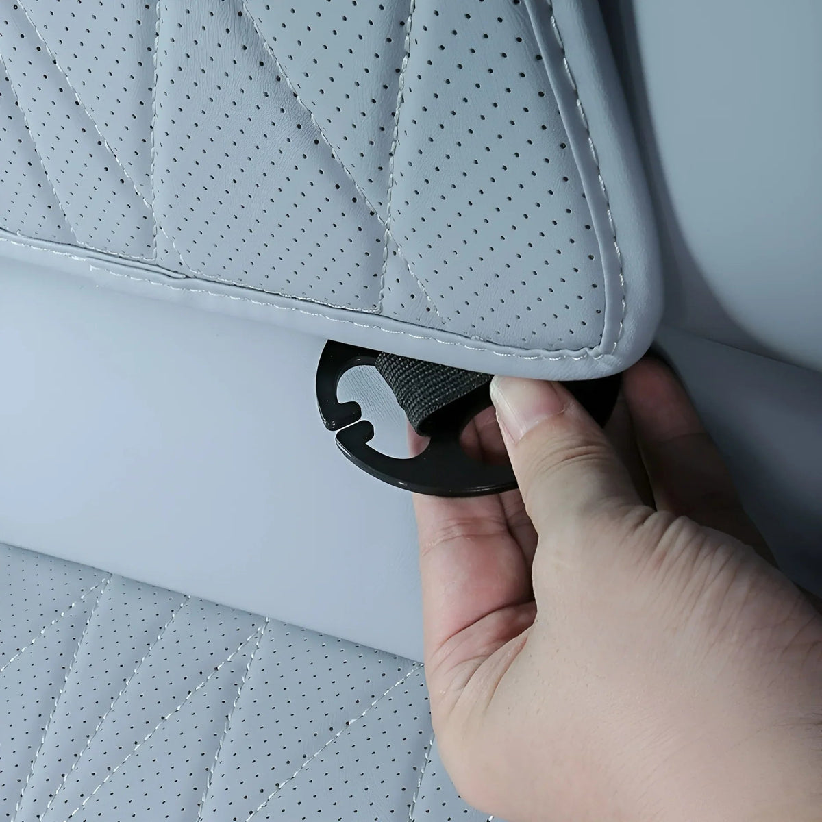 Close-up of a hand attaching a black strap to a perforated grey leather car seat cover.