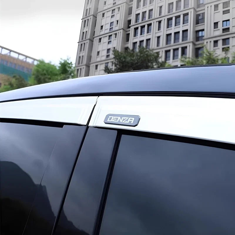 Close-up of a silver DENZA emblem on a sleek black vehicle roofline against an urban building background.